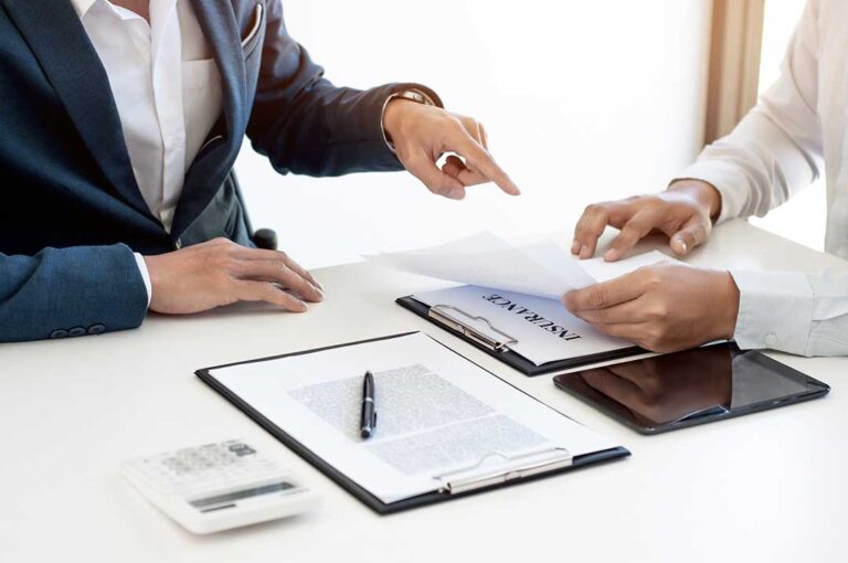A person reviews health insurance paperwork at a kitchen table, with forms, a pen, and a cup of coffee visible.